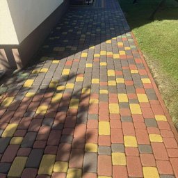 BŁYSK - Kolorowa kostka brukowa w odcieniach czerwieni, żółci i szarości, tworząca wzór na ścieżce obok trawnika. Część ścieżki zacieniona, widoczne schody i huśtawka w tle.