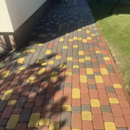 Kolorowa kostka brukowa w odcieniach czerwieni, ż&oacute;łci i szarości, tworząca wz&oacute;r na ścieżce obok trawnika. Część ścieżki zacieniona, widoczne schody i huśtawka w tle.