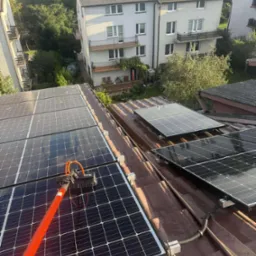 Czyszczenie paneli fotowoltaicznych na dachu domu za pomocą szczotki na teleskopowym kiju. W tle budynki mieszkalne i zieleń.