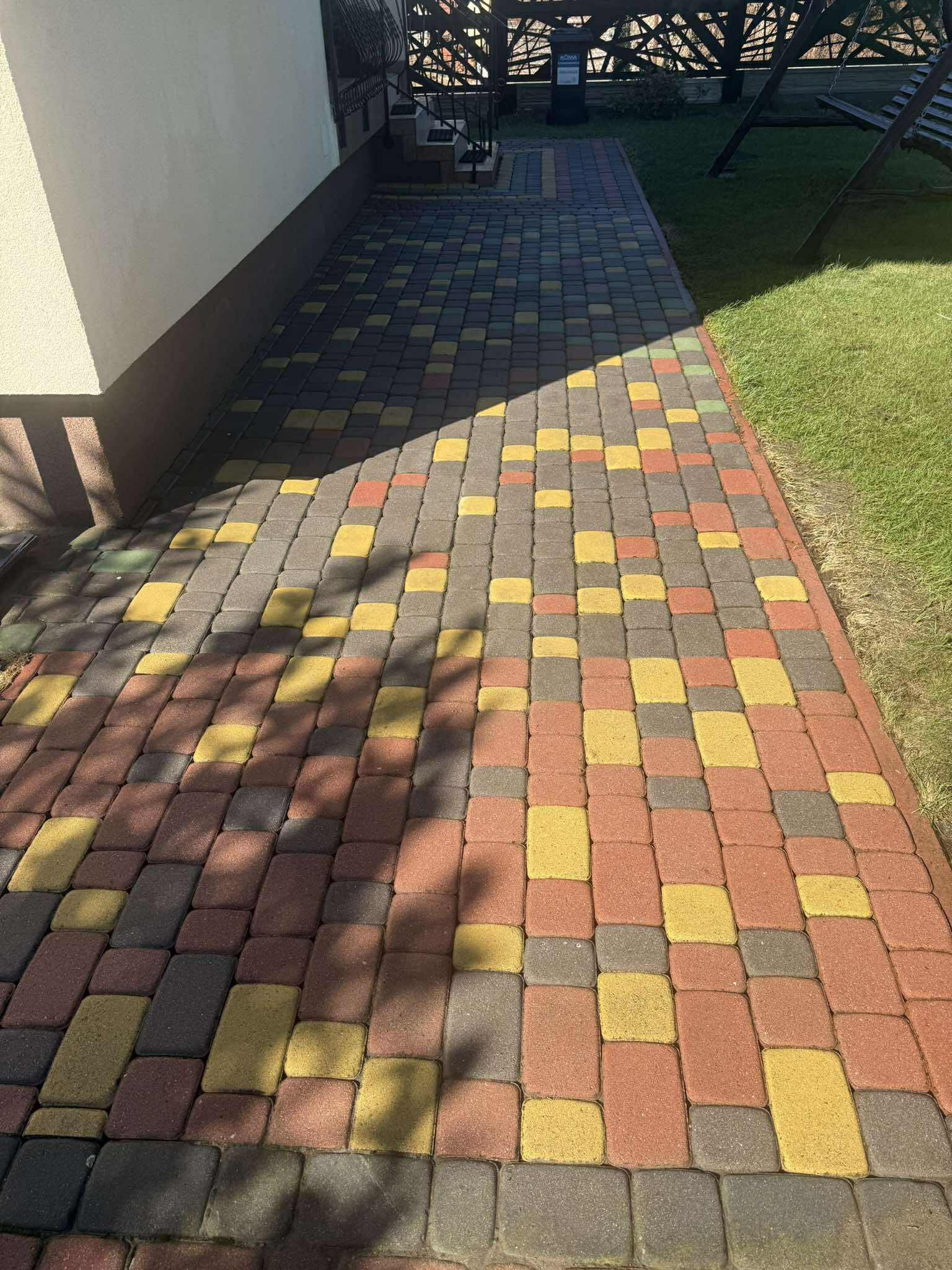Kolorowa kostka brukowa w odcieniach czerwieni, żółci i szarości, tworząca wzór na ścieżce obok trawnika. Część ścieżki zacieniona, widoczne schody i huśtawka w tle.