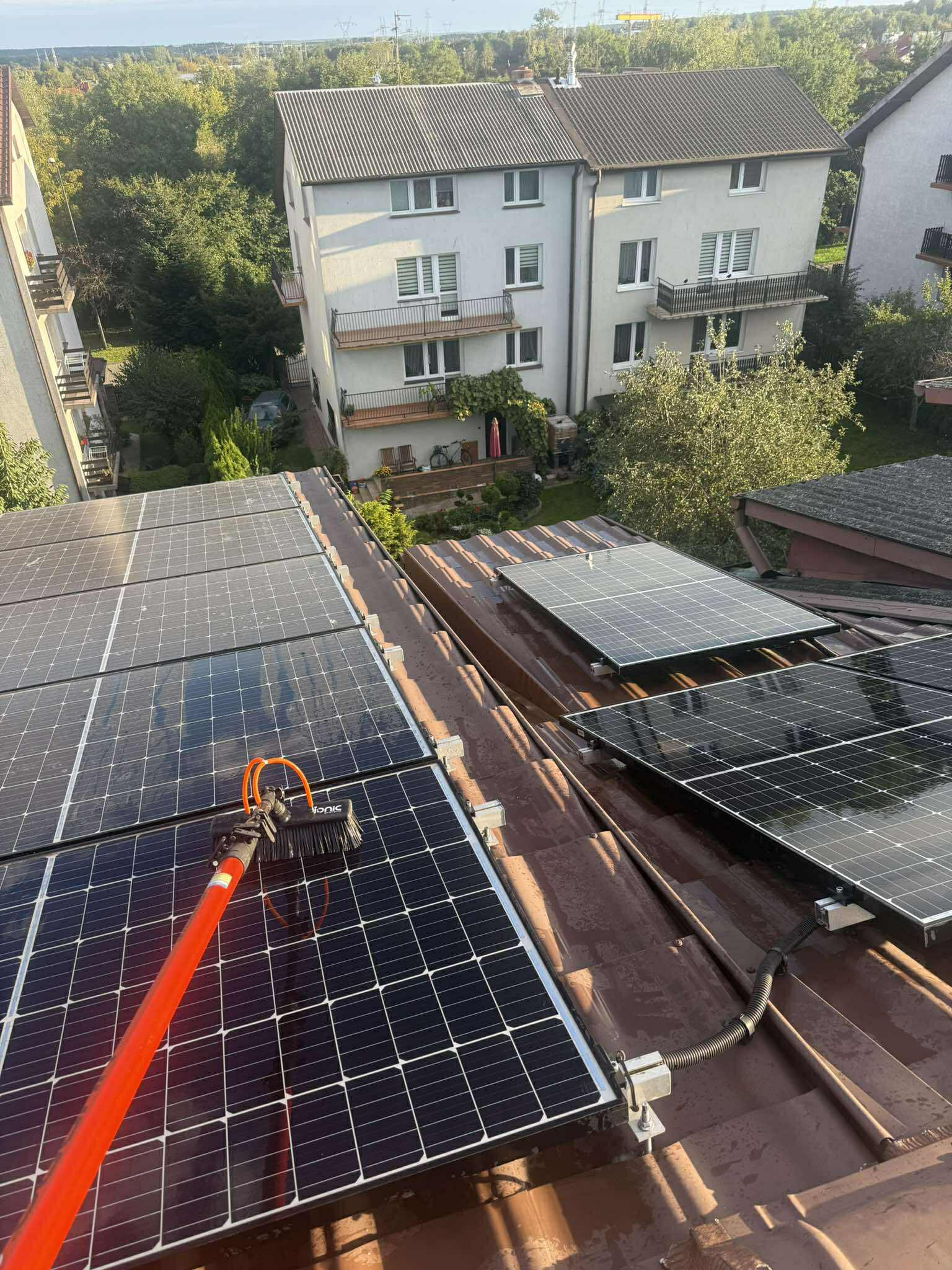 Czyszczenie paneli fotowoltaicznych na dachu domu za pomocą szczotki na teleskopowym kiju. W tle budynki mieszkalne i zieleń.