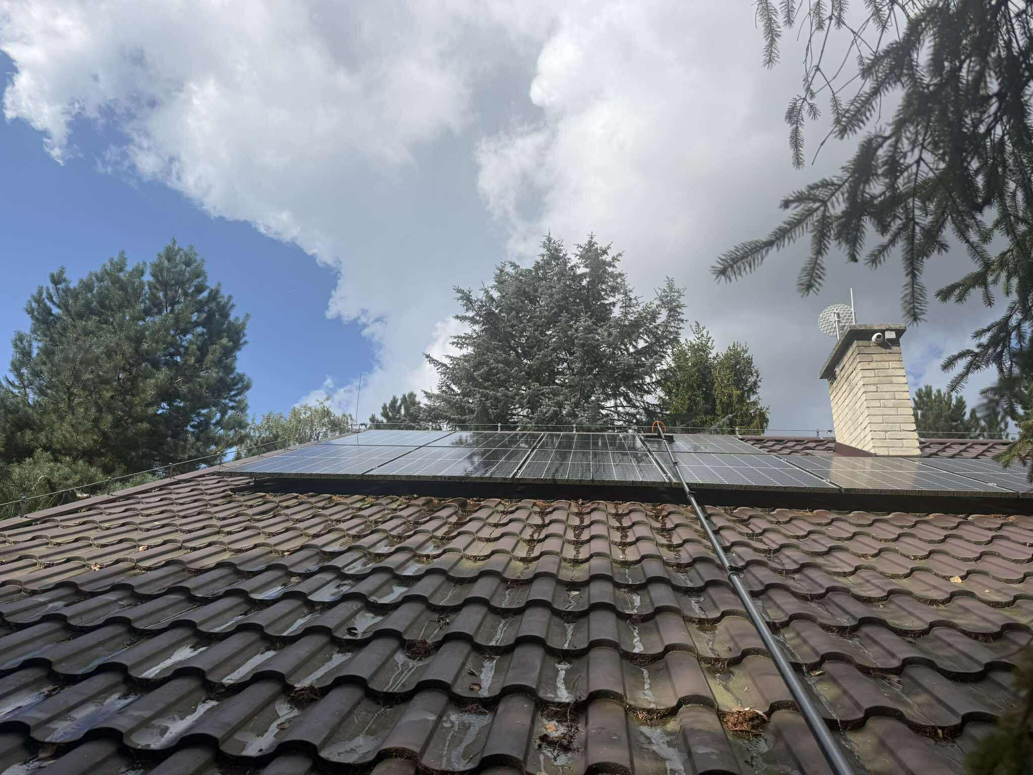 Czyszczenie paneli słonecznych na brązowym dachu z użyciem długiej szczotki teleskopowej. Widoczne drzewa i pochmurne niebo w tle. Ujęcie z dołu.