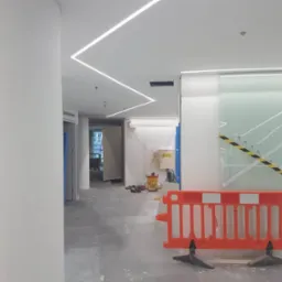 Wnętrze biura w trakcie wykończenia: białe ściany, kolumna, sufit z liniowym oświetleniem LED, pomarańczowa barierka zabezpieczająca i narzędzia w tle.