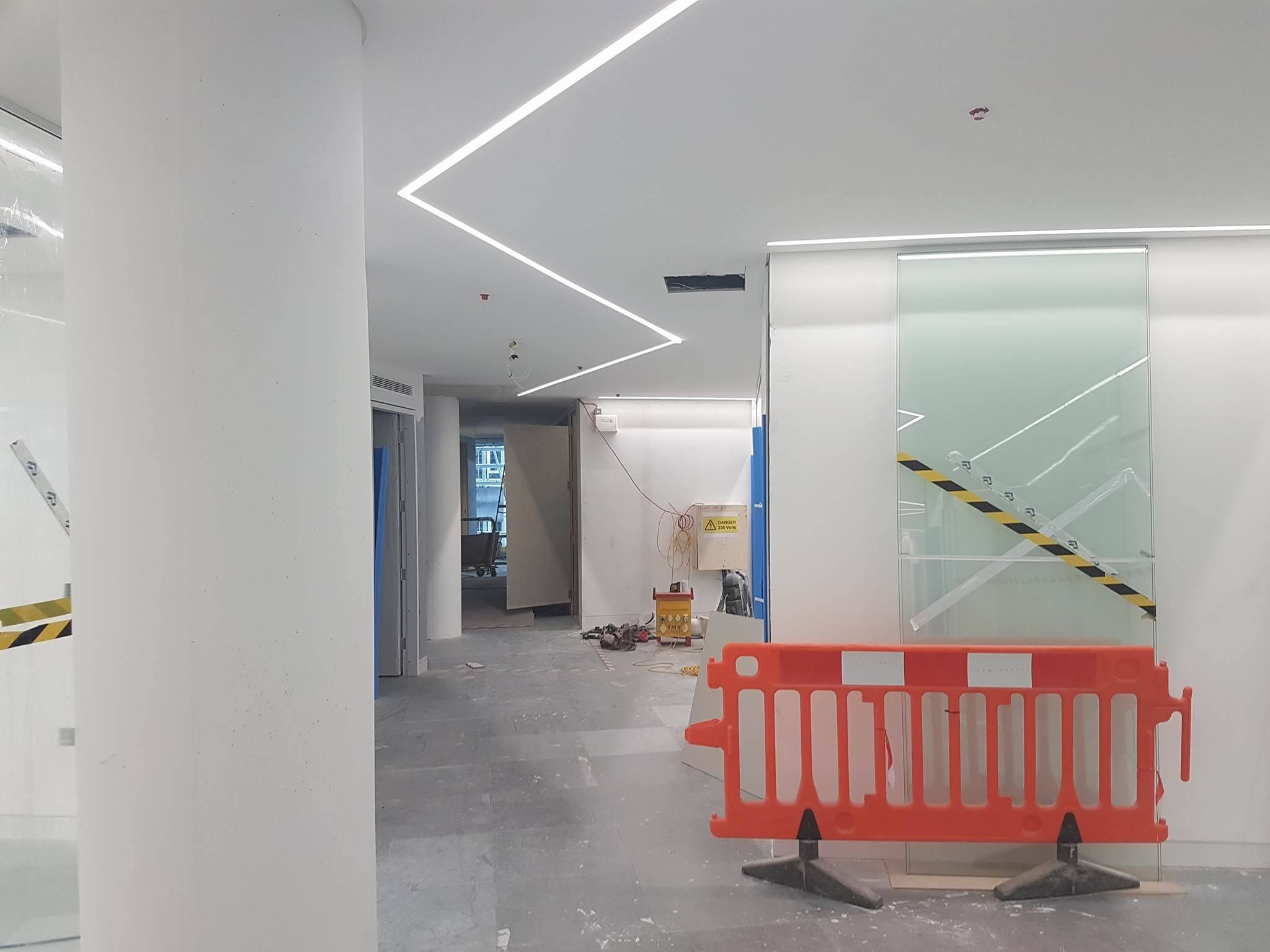 Wnętrze biura w trakcie wykończenia: białe ściany, kolumna, sufit z liniowym oświetleniem LED, pomarańczowa barierka zabezpieczająca i narzędzia w tle.