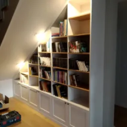 Biała zabudowa pod schodami z półkami z jasnego drewna, wypełniona książkami. Widoczne oświetlenie punktowe i skrzynka z narzędziami na podłodze.