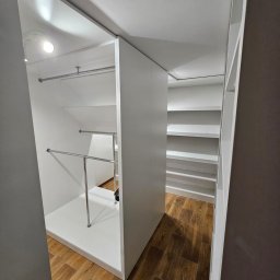 VERTIKA S.C. - Nowoczesna, biała garderoba z drążkami i półkami, widok z perspektywy wejścia. Jasne oświetlenie podkreśla minimalistyczny design i drewnianą podłogę.