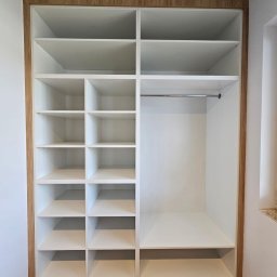 VERTIKA S.C. - Wnętrze białej szafy wnękowej z drewnianym obramowaniem, widok na półki i drążek na ubrania, podłoga z paneli imitujących drewno.