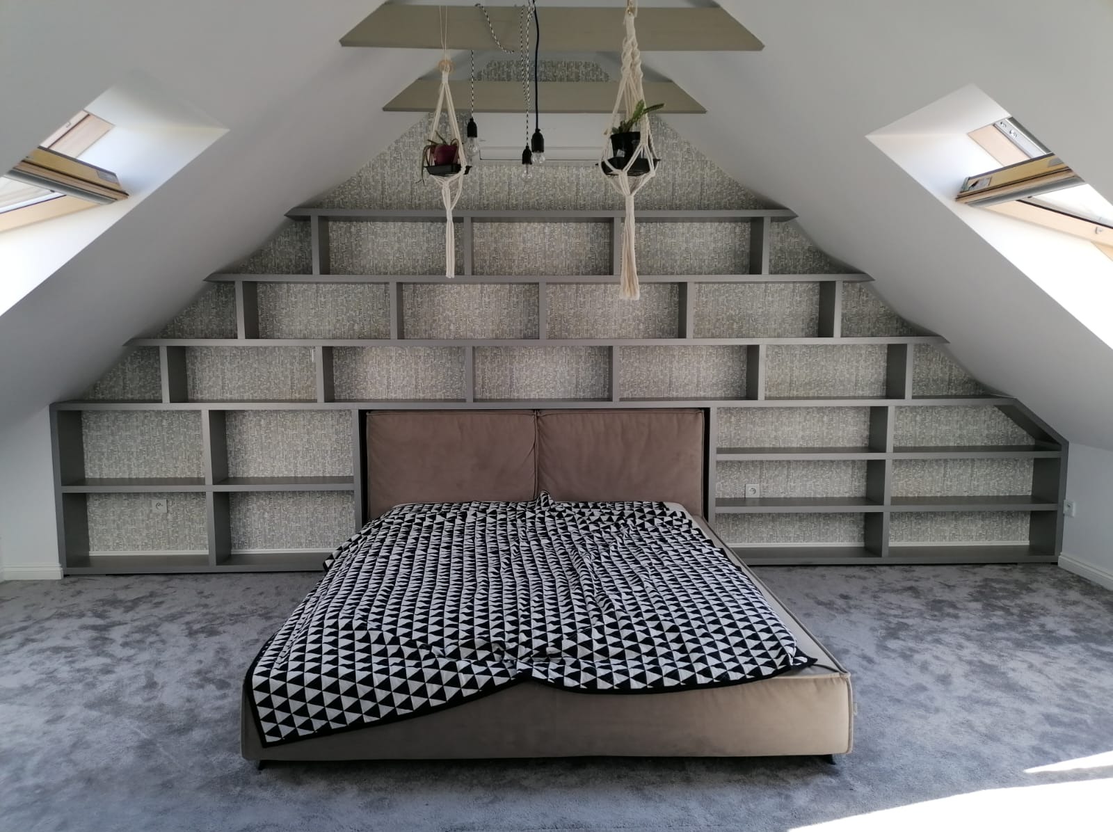 Pokój na poddaszu z łóżkiem i regałem w kształcie schodów, wbudowanym w ścianę. Rośliny wiszące pod sufitem. Ujęcie z dołu, podkreślające przestrzeń i geometrię.