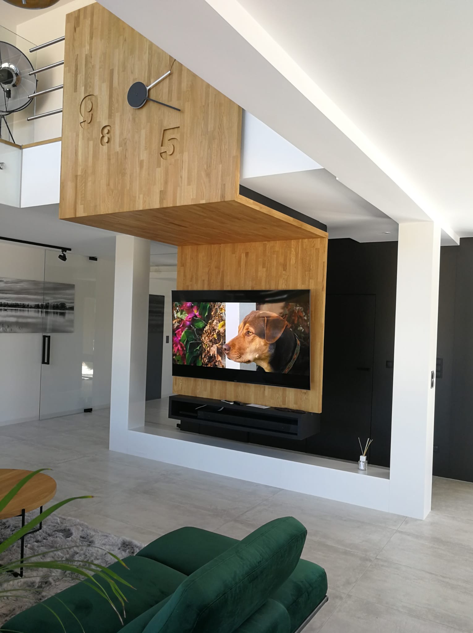 Nowoczesny salon z drewnianym zegarem ściennym i telewizorem z obrazem psa. Minimalistyczny design, jasne kolory i naturalne materiały tworzą przytulną przestrzeń.