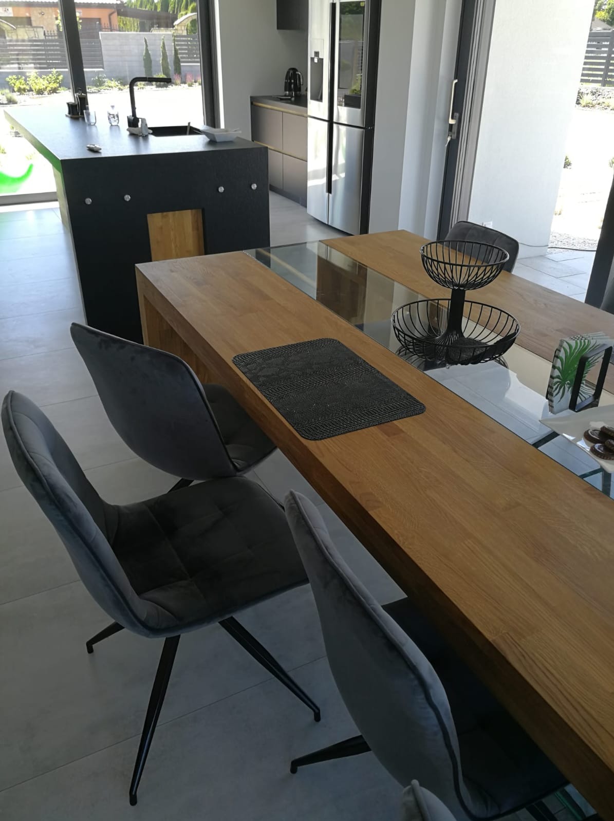 Nowoczesna kuchnia z wyspą i drewnianym stołem z szarymi krzesłami. Minimalistyczny design, jasne wnętrze z dużymi oknami i widokiem na zieleń.