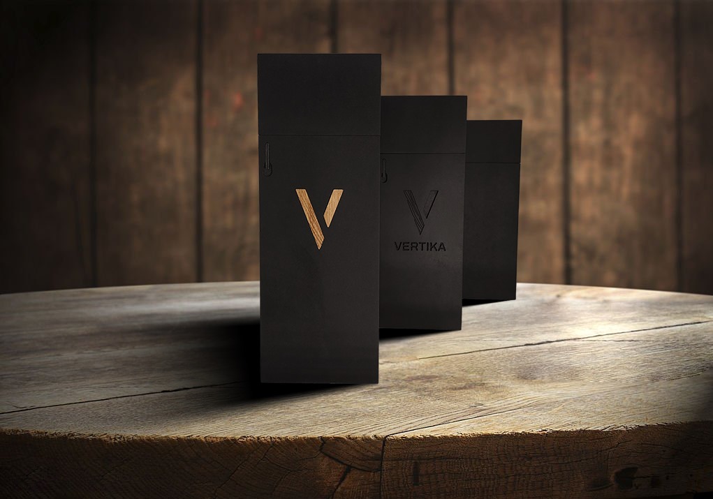 Trzy czarne, eleganckie opakowania na drewnianym blacie, z logo 'V' w kolorze drewna na pierwszym planie, na tle drewnianej ściany. Minimalistyczny design.