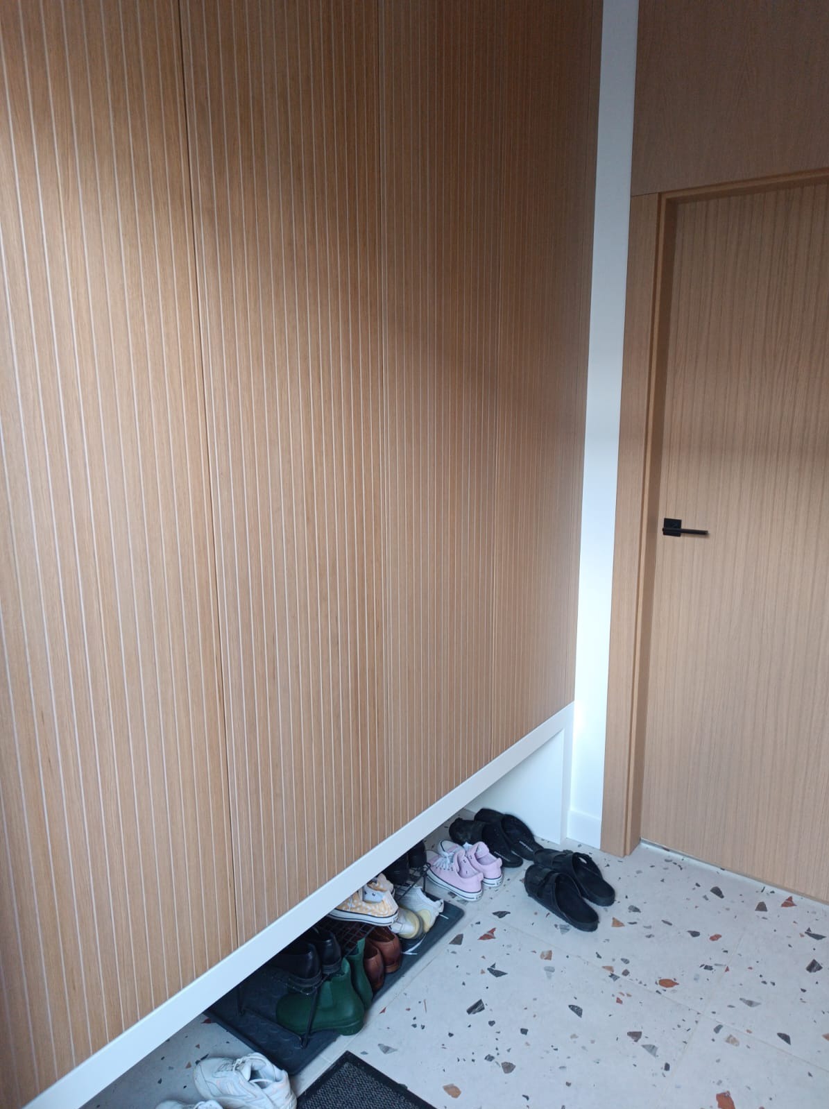 Szafa wnękowa z drewnianymi lamelami i miejscem na buty, z widocznymi butami na podłodze z terazzo w przedpokoju. Minimalistyczny design.