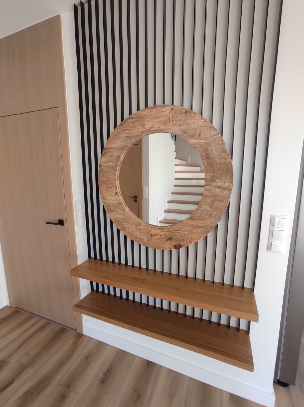 Drewniane lustro w okrągłej ramie i półki z drewna w przedpokoju. W tle widoczne schody i pionowe, czarne listwy dekoracyjne na ścianie.