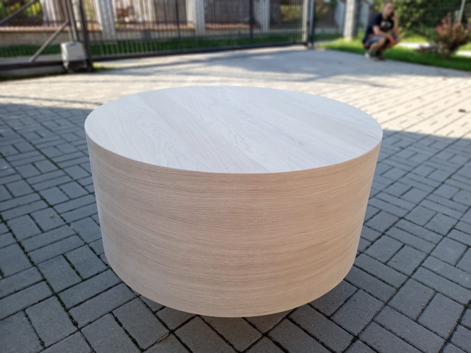 Okrągły, niski stolik z jasnego drewna na kostce brukowej. W tle metalowa brama i osoba. Widoczne delikatne usłojenie drewna.