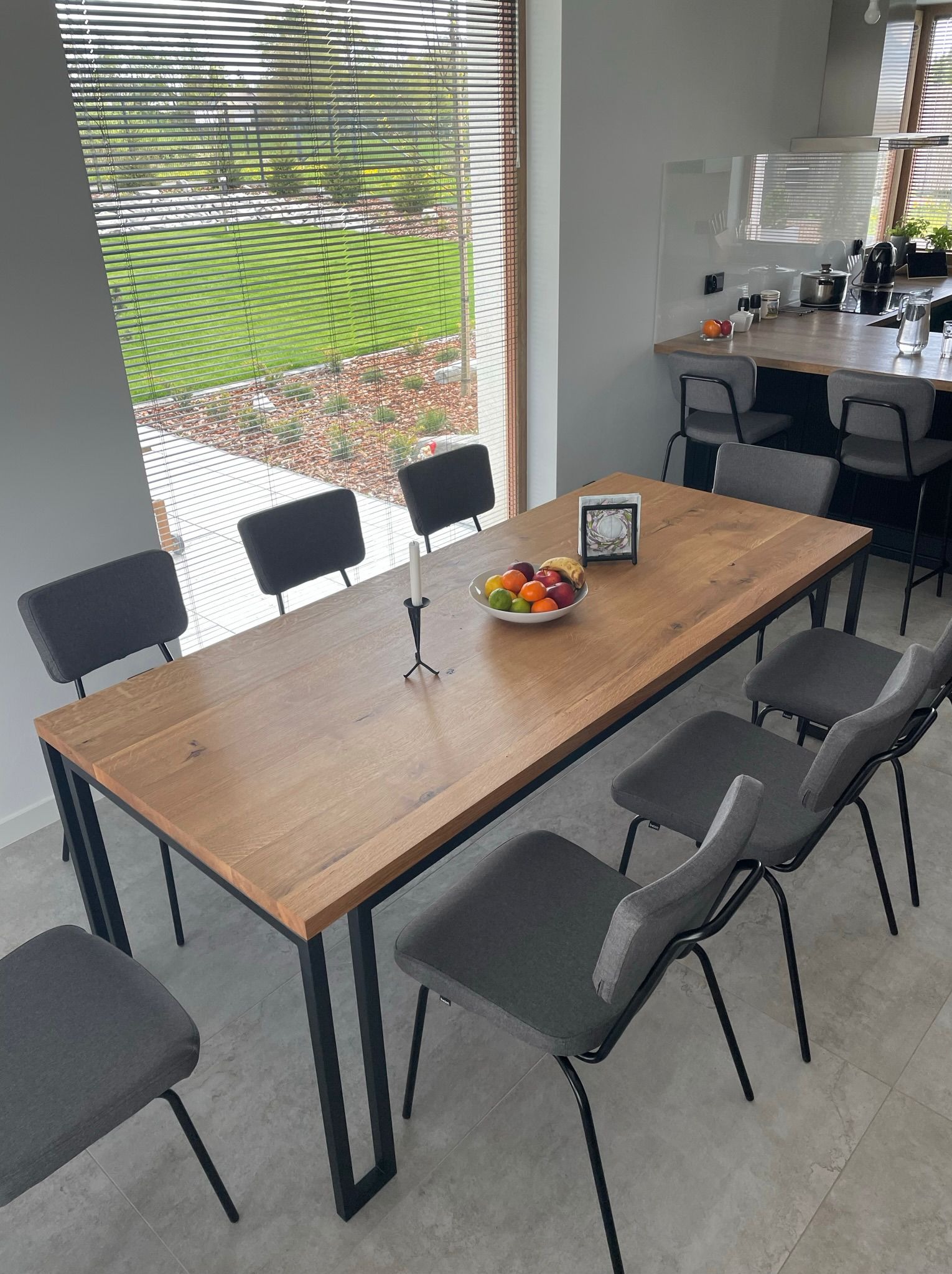 Stół z blatem z jasnego drewna i czarnymi nogami, otoczony szarymi krzesłami w nowoczesnej jadalni z widokiem na zieleń przez żaluzje okienne.