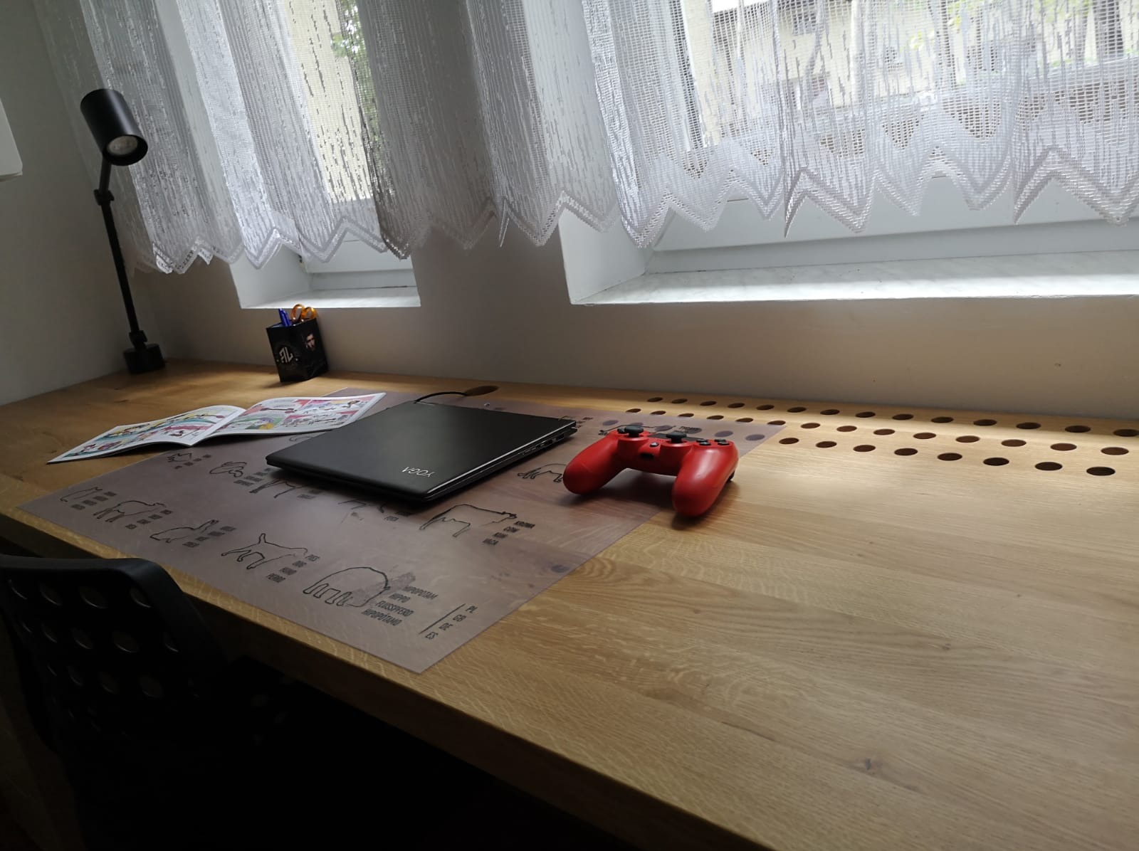 Drewniany blat biurka z laptopem, gamepadem i lampką. W tle okno z firanką. Widoczny fragment krzesła. Blat z otworami wentylacyjnymi przy ścianie.