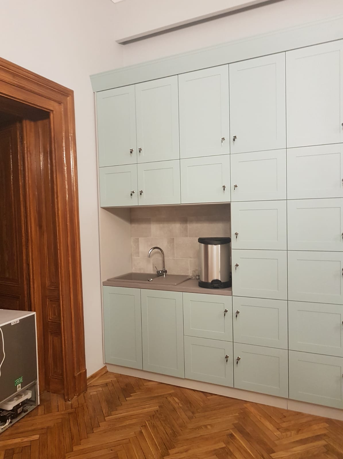 Jasnoniebieska zabudowa kuchenna na wymiar z licznymi szafkami, srebrnym zlewem i dystrybutorem wody, na tle parkietu w jodełkę i drewnianych drzwi.