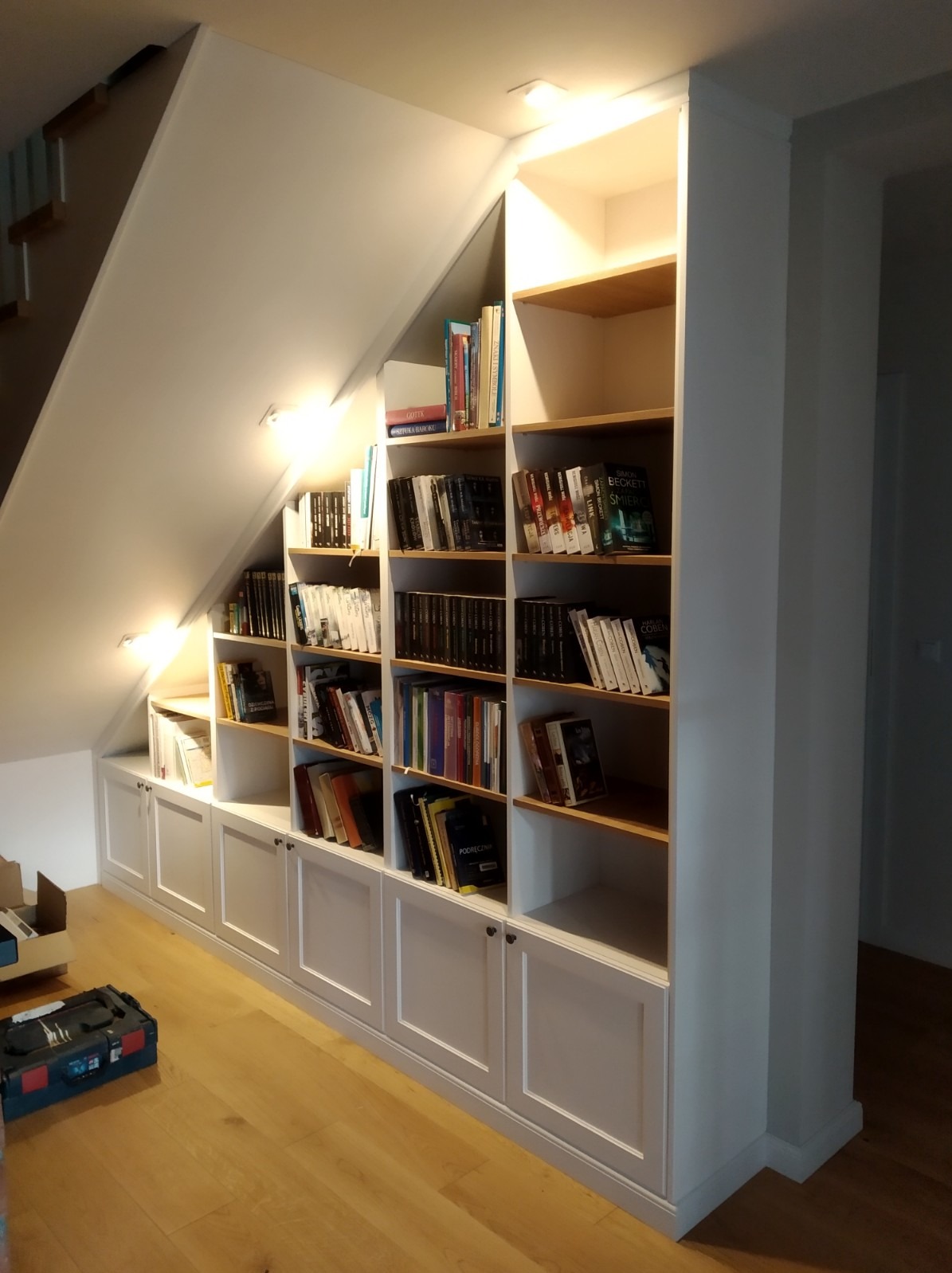 Biała zabudowa pod schodami z półkami z jasnego drewna, wypełniona książkami. Widoczne oświetlenie punktowe i skrzynka z narzędziami na podłodze.