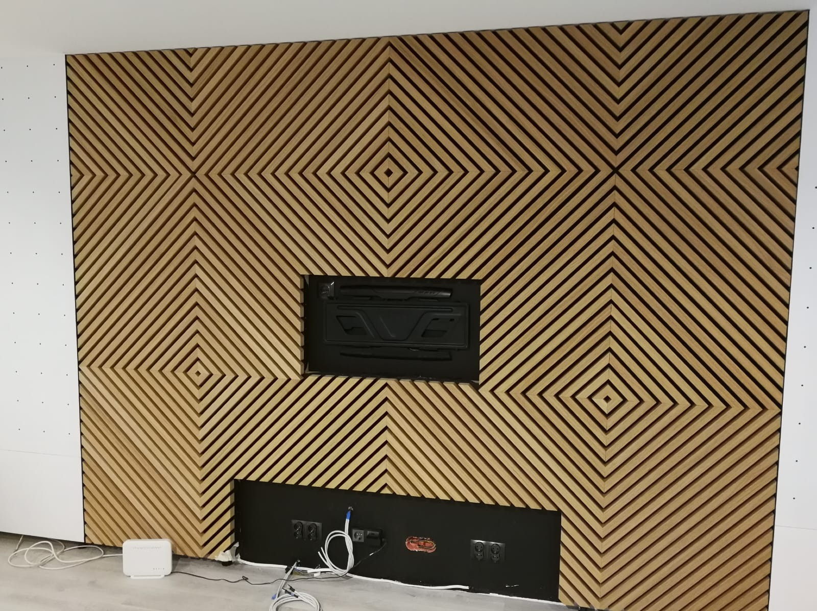 Dekoracyjna ściana z drewnianych listew ułożonych w geometryczny wzór rombów. Widoczne otwory na kable i mocowania, białe panele po bokach. Nowoczesny design.