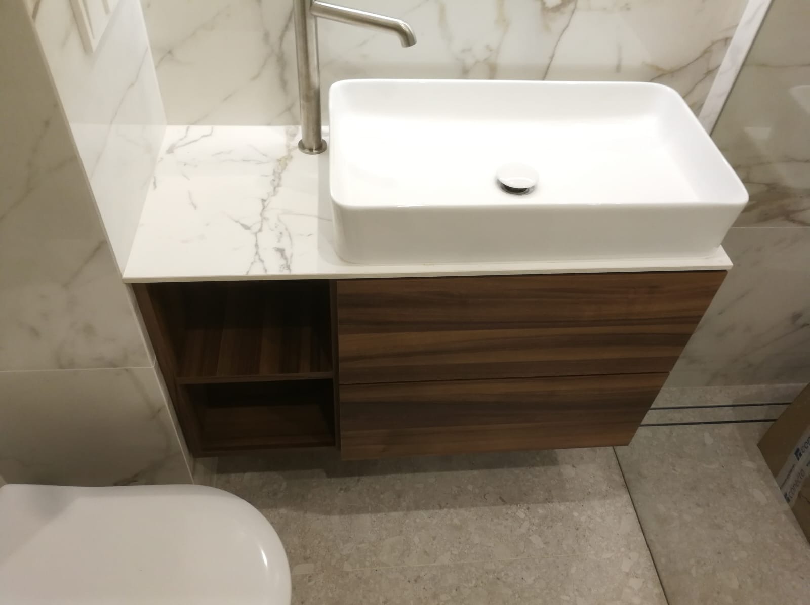 Nowoczesna łazienka z białą, prostokątną umywalką nablatową i drewnianą szafką z otwartą półką. Marmurowe ściany i podłoga w odcieniach szarości.