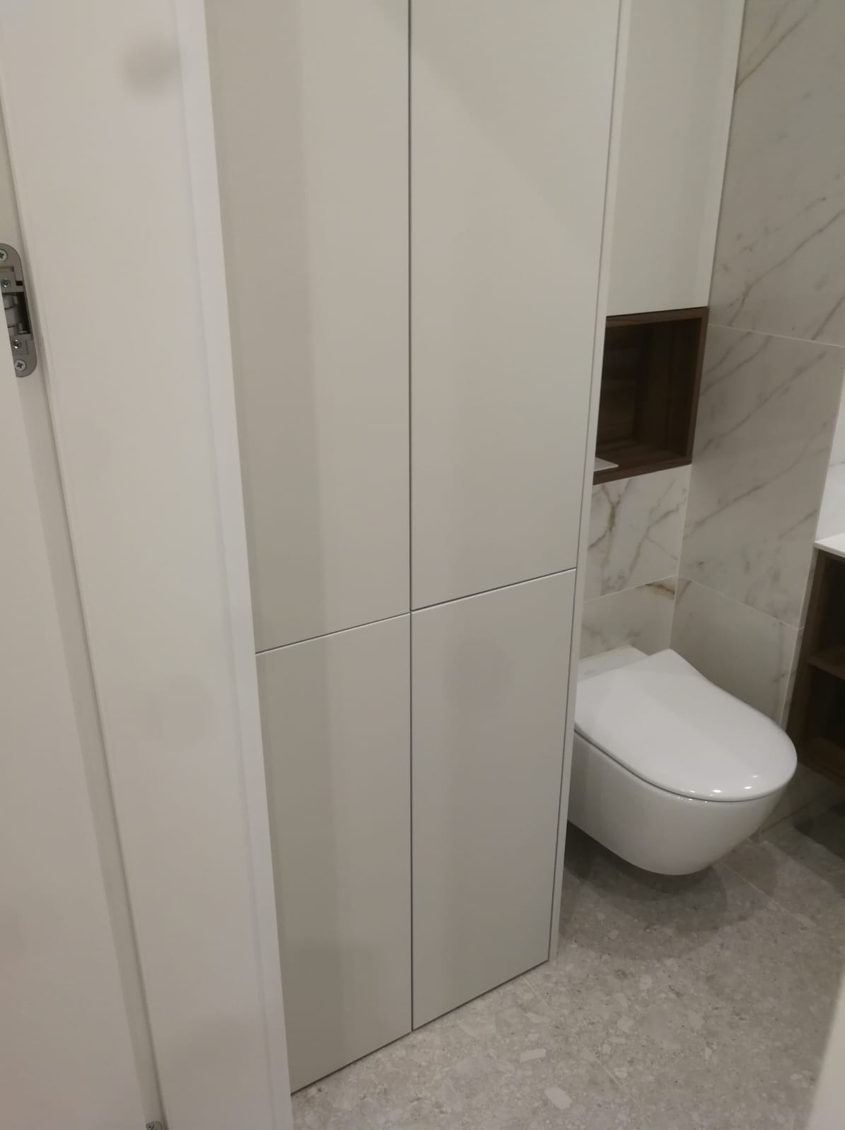 Biała szafa wnękowa z półkami w łazience z marmurowymi płytkami i podwieszaną toaletą. Minimalistyczny design, nowoczesne wykończenie.