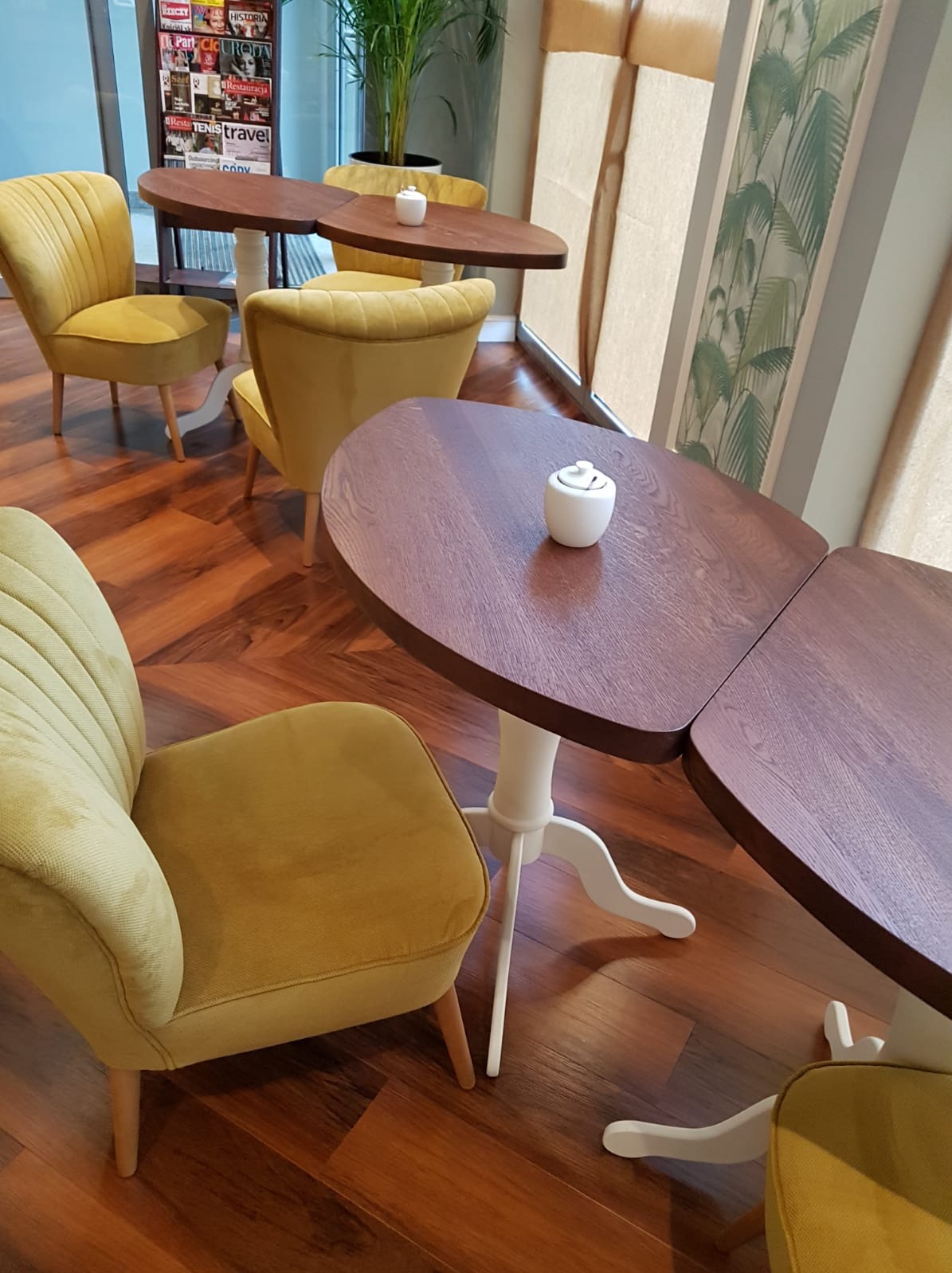 Drewniane blaty stolików kawowych o nieregularnym kształcie, wsparte na białych nogach, z żółtymi krzesłami na parkiecie w jodełkę, w kawiarni.