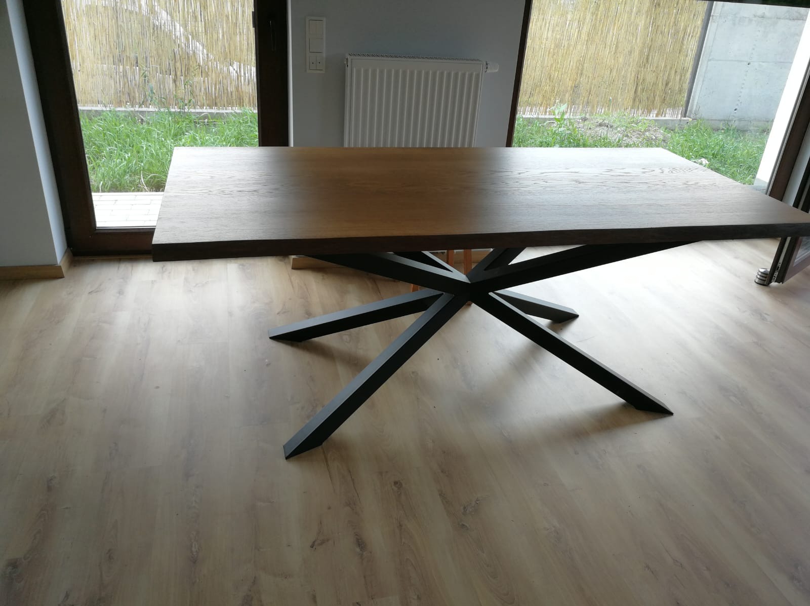 Stół z blatem z jasnego drewna i czarną, geometryczną podstawą na tle okna i grzejnika, na podłodze z paneli. Widok z perspektywy podłogi.