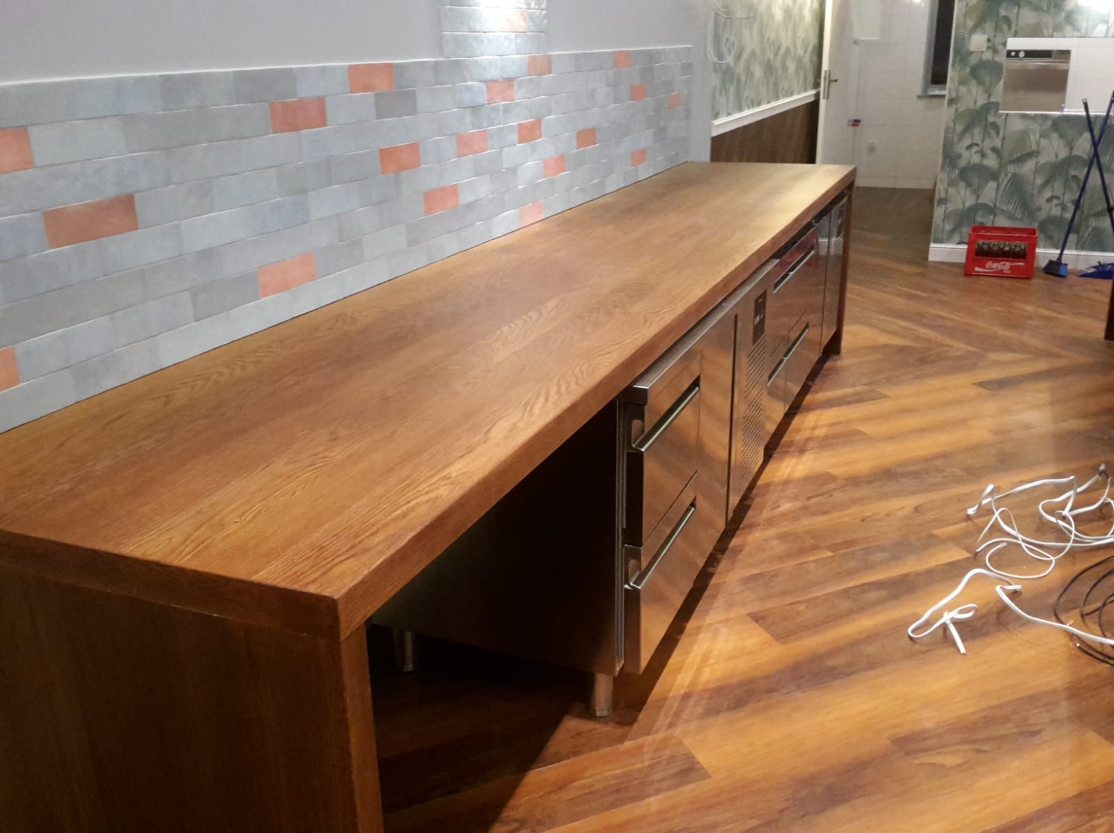 Długi, drewniany blat z szafkami ze stali nierdzewnej, umieszczony pod ścianą z dekoracyjnymi płytkami. Widoczne elementy wyposażenia kuchni i podłoga z paneli.