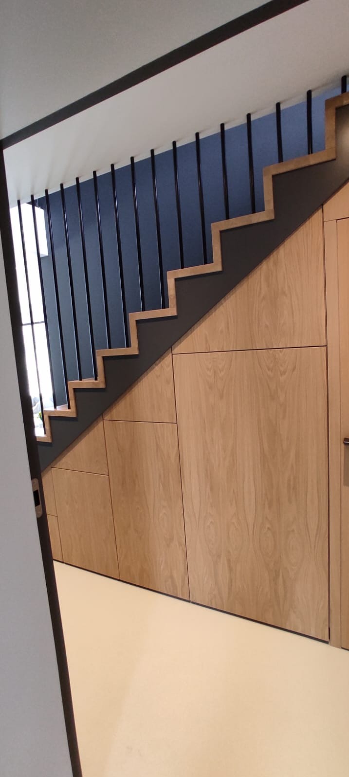 Schody z zabudową drewnianą, czarne barierki na tle błękitnej ściany. Naturalne wykończenie drewna i nowoczesny design.