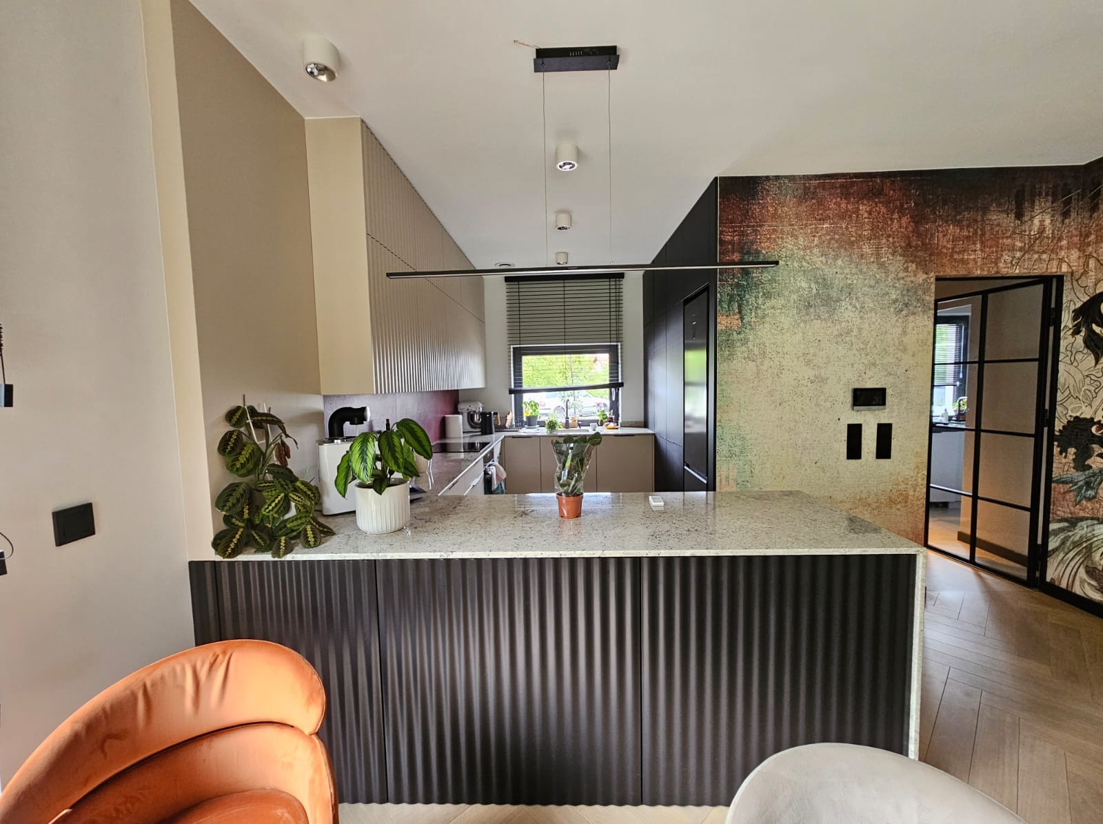 Nowoczesna kuchnia z wyspą i blatem z jasnego kamienia, fronty szafek z ciemnego, ryflowanego drewna. Minimalistyczne oświetlenie i rośliny doniczkowe.