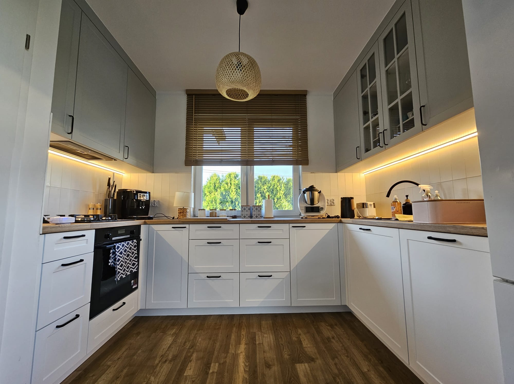 Nowoczesna kuchnia na wymiar w Wieliczce. Białe fronty dolne, szare górne szafki. Drewniany blat i podłoga. Oświetlenie LED pod szafkami.