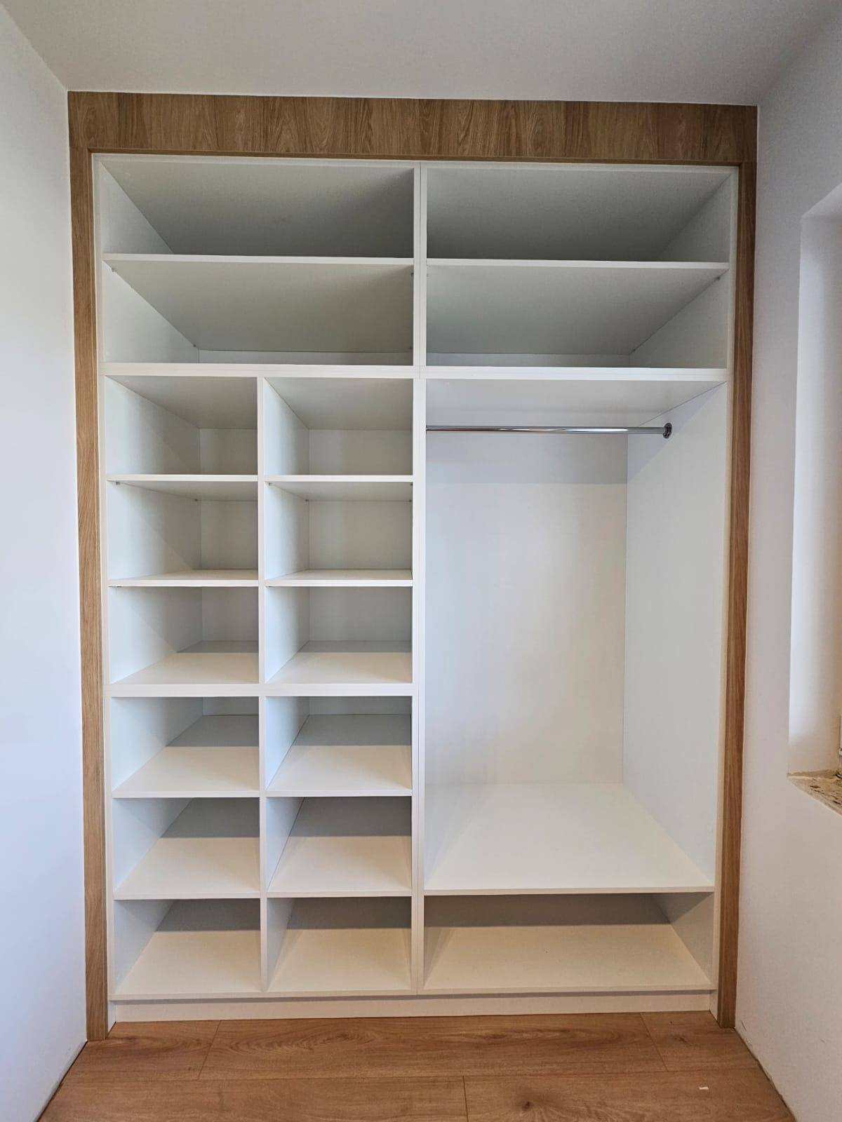 Wnętrze białej szafy wnękowej z drewnianym obramowaniem, widok na półki i drążek na ubrania, podłoga z paneli imitujących drewno.