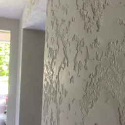 Dekoracyjna ściana z wyraźną fakturą w odcieniach szarości, widoczny fragment przejścia do innego pomieszczenia. Specyficzny wzór na ścianie, światło dzienne.