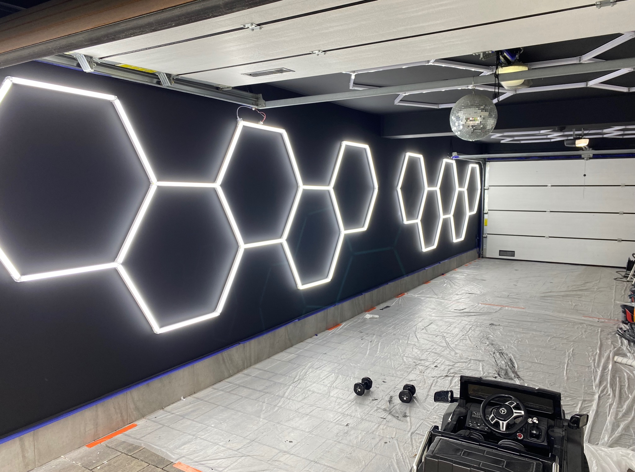 Czarna ściana z sześciokątnymi lampami LED w garażu, zabezpieczona folią podłoga, widoczny fragment zabawkowego samochodu i dyskotekowa kula.