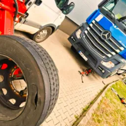 Wymiana opony ciężarówki z użyciem specjalistycznego sprzętu. Widoczny duży opona, ciężarówka Mercedes i biały bus w tle. Oświetlenie dzienne.