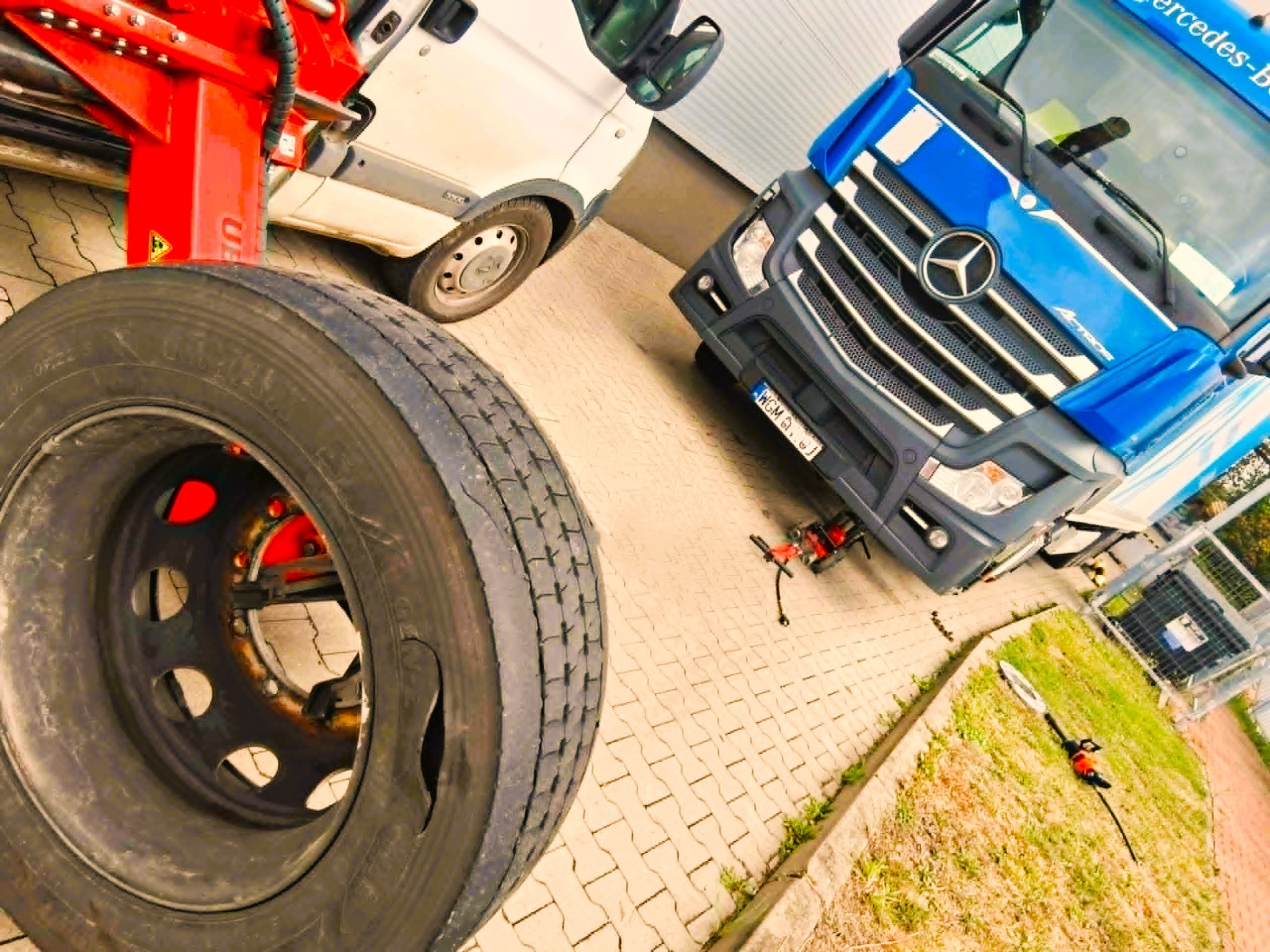 Wymiana opony ciężarówki z użyciem specjalistycznego sprzętu. Widoczny duży opona, ciężarówka Mercedes i biały bus w tle. Oświetlenie dzienne.