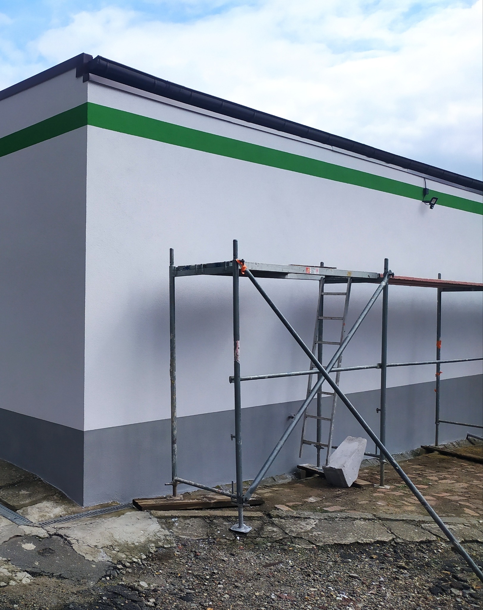 Świeżo pomalowana ściana budynku z szarą lamperią i zielonym pasem. Widoczne rusztowanie i drabina, sugerujące prace wykończeniowe.