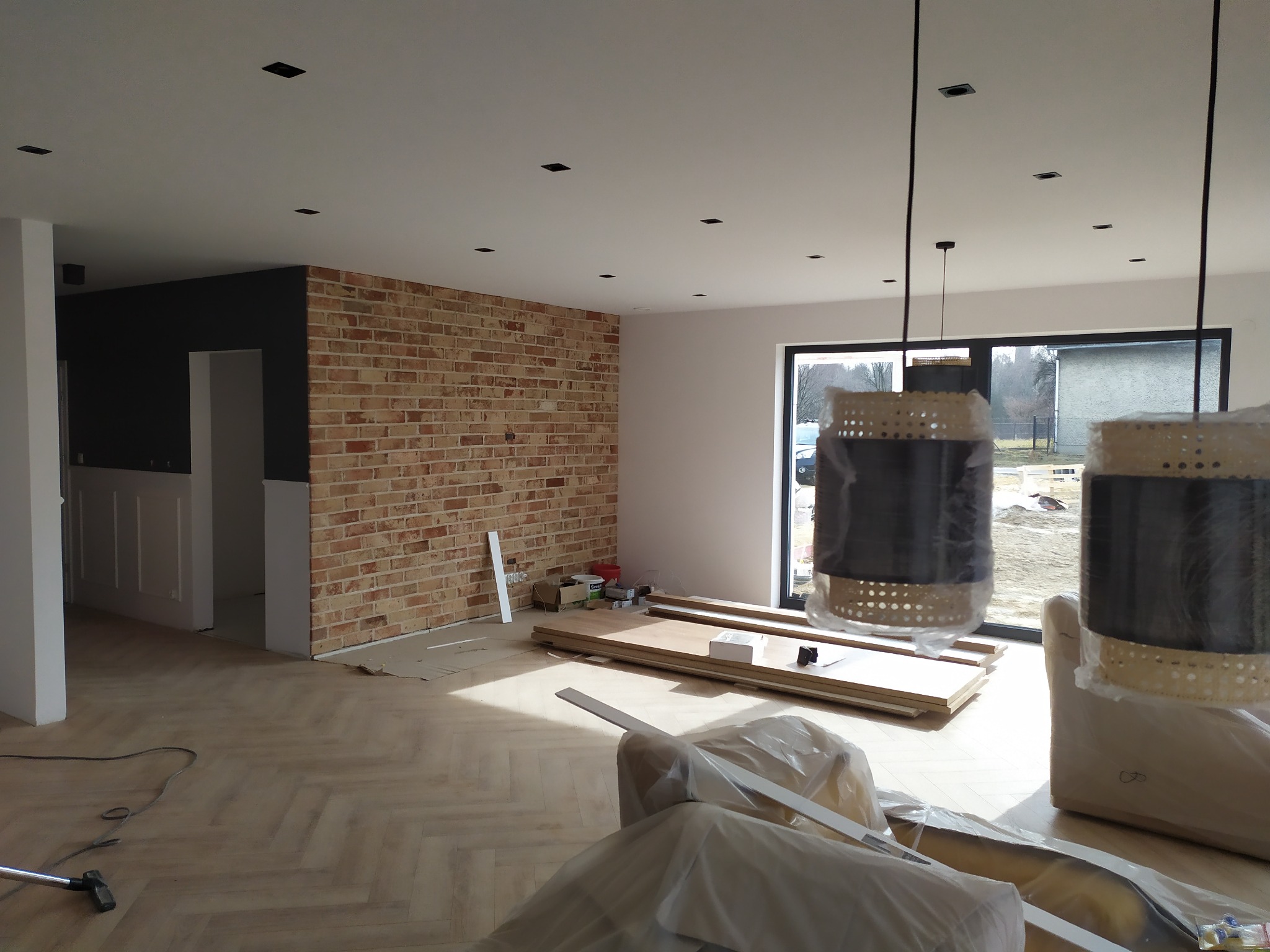 Jasne parkiet w jodełkę, ceglana ściana z czarną wstawką, widoczne elementy wykończeniowe i lampy w folii, duże okno. Wnętrze w trakcie prac wykończeniowych.