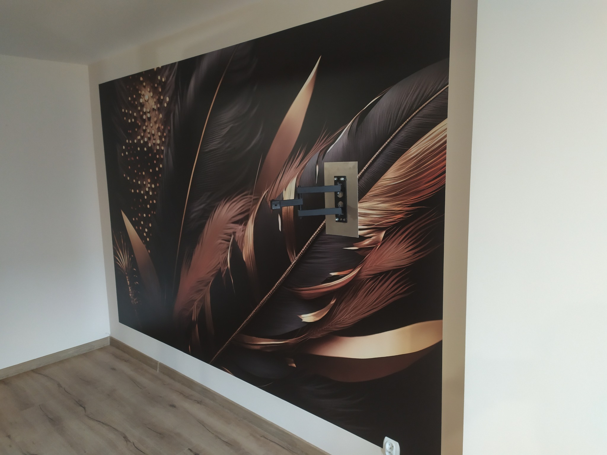 Ściana z fototapetą z motywem piór w odcieniach brązu i czerni, z zamontowanym uchwytem na telewizor. Widoczna podłoga w kolorze drewna i białe ściany.