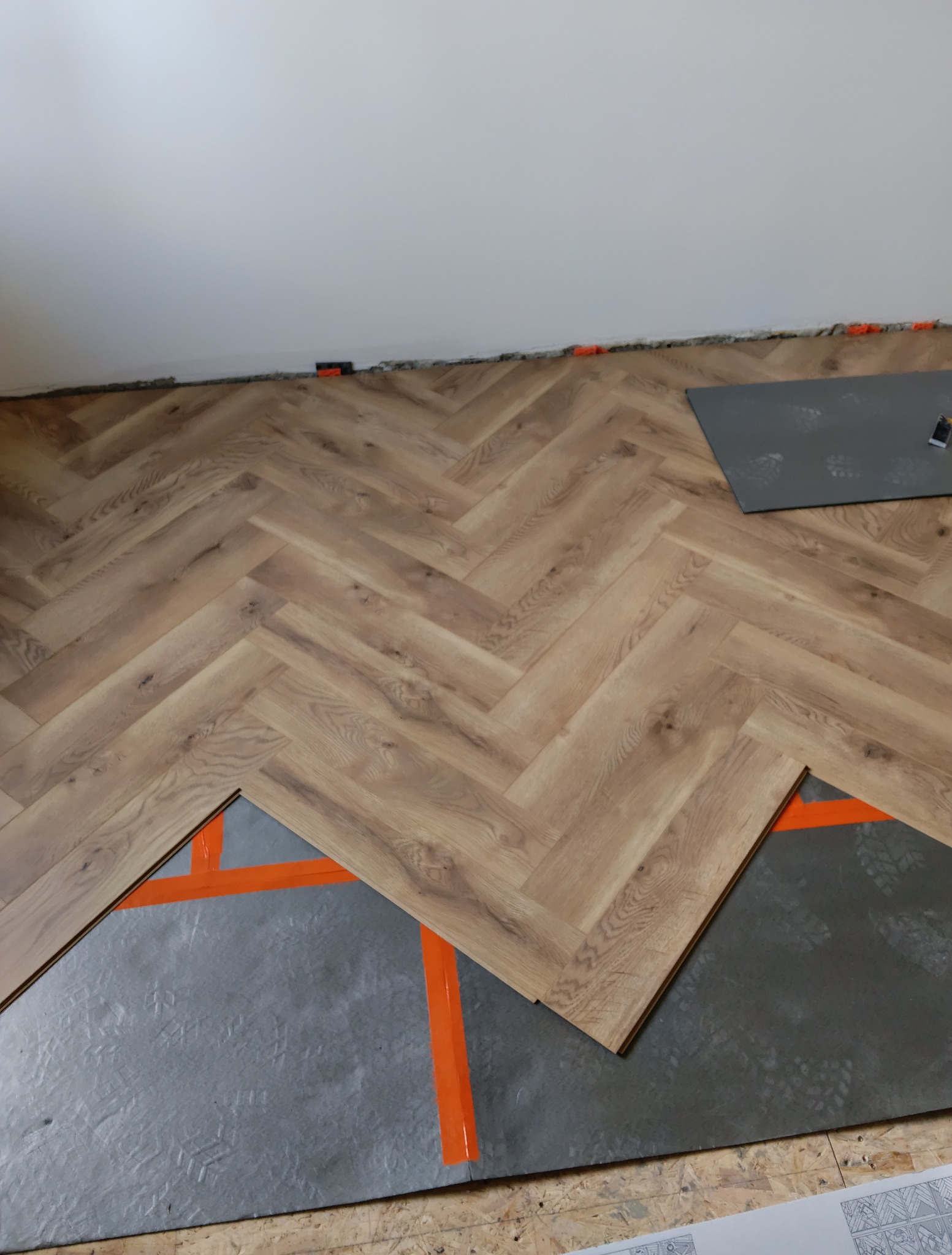 Układanie paneli podłogowych w jodełkę na podkładzie z pomarańczową taśmą. Widoczne panele, podkład i fragment ściany. Etap prac wykończeniowych.