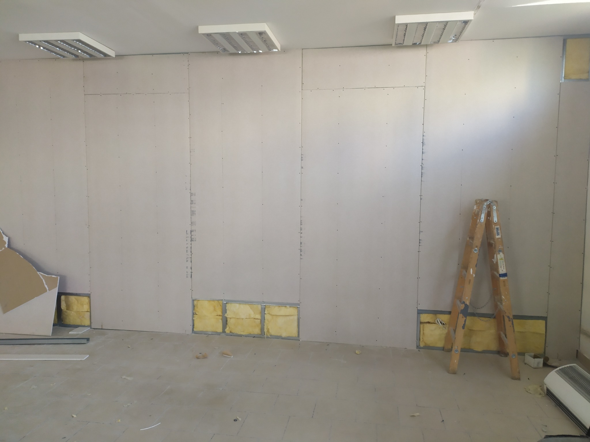 Montaż płyty karton gips: ściana z widoczną izolacją, drabina oparta o ścianę, surowe wykończenie podłoża, oświetlenie sufitowe.