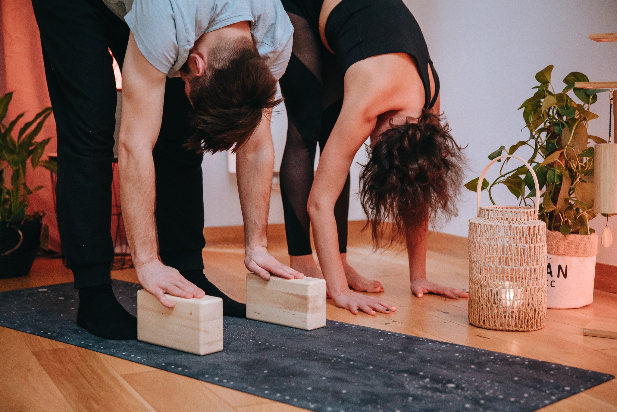 Dwie osoby w skłonie podczas zajęć jogi, używające drewnianych klocków na macie. W tle rośliny doniczkowe i lampion, ciepłe oświetlenie tworzy relaksującą atmosferę.