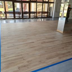 Tomi Parkiety - Jasna podłoga z paneli drewnopodobnych, oddzielona niebieską taśmą od szarych płytek. W tle widoczne okna i odbicie osoby w lustrzanej kolumnie.