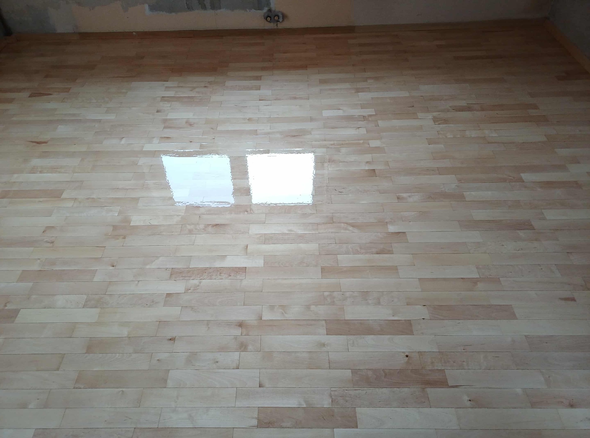 Jasny parkiet z wyraźnym lakierem odbijającym światło okien. Widoczne niedoskonałości sufitu w tle. Świeżo położona podłoga.