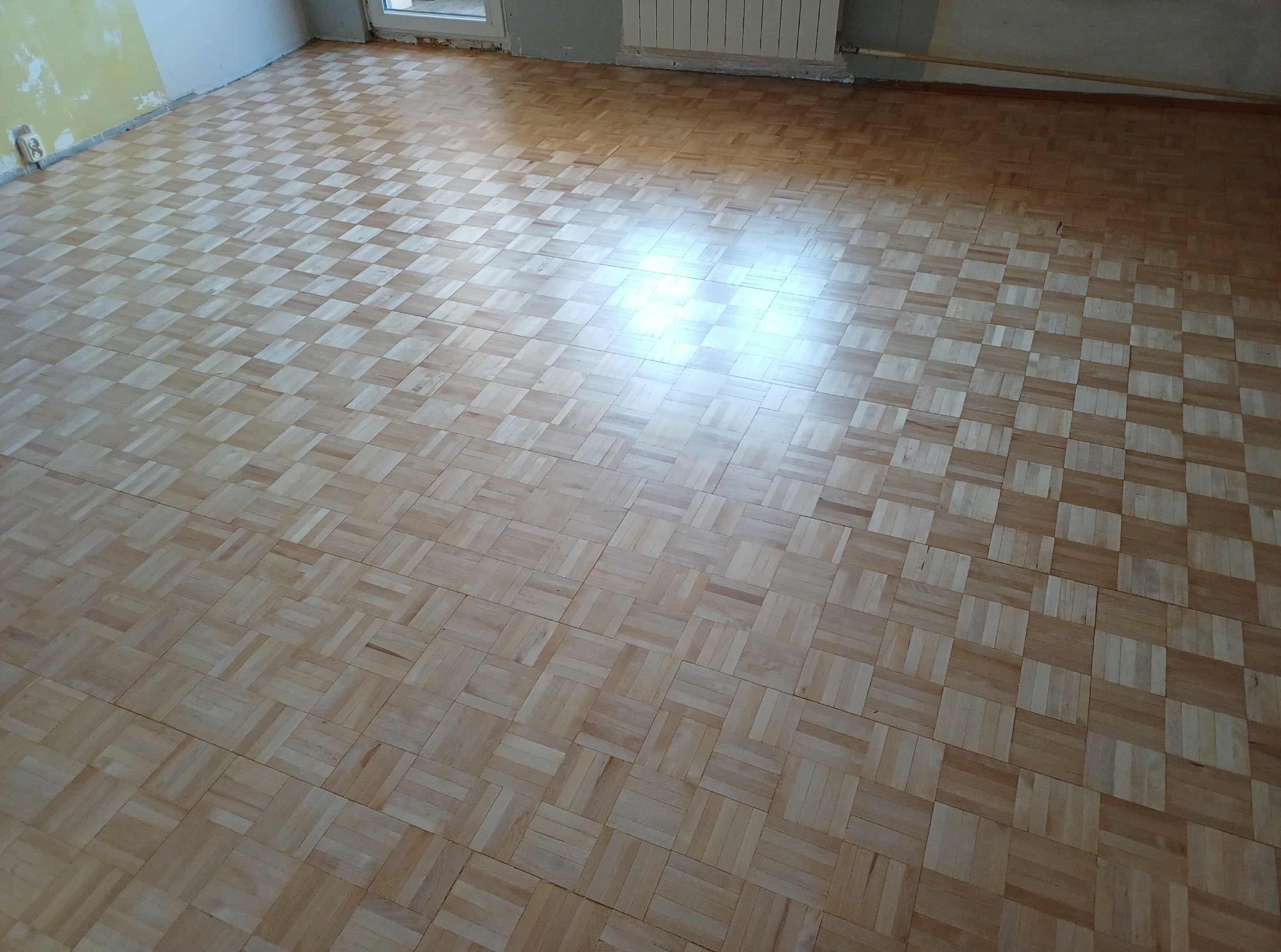 Ukończona podłoga z mozaiki parkietowej w jasnym odcieniu drewna, ułożona w pomieszczeniu z fragmentami ścian przygotowanych do malowania. Widoczne okno i grzejnik.