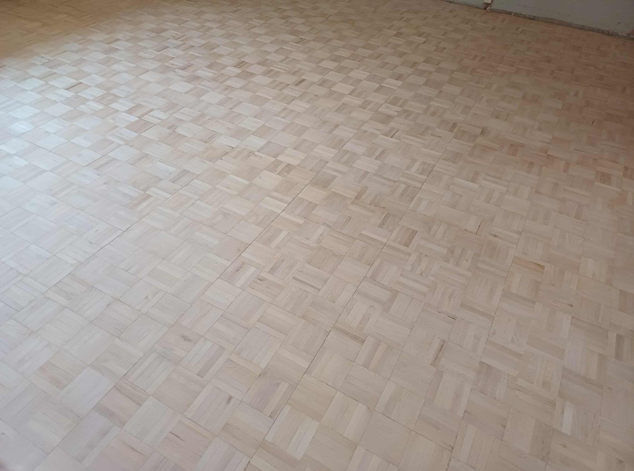 Jasna, parkietowa podłoga ułożona w kwadraty, widziana z perspektywy. Świeżo położony parkiet z wyraźnym wzorem klepek. W tle widoczna ściana.