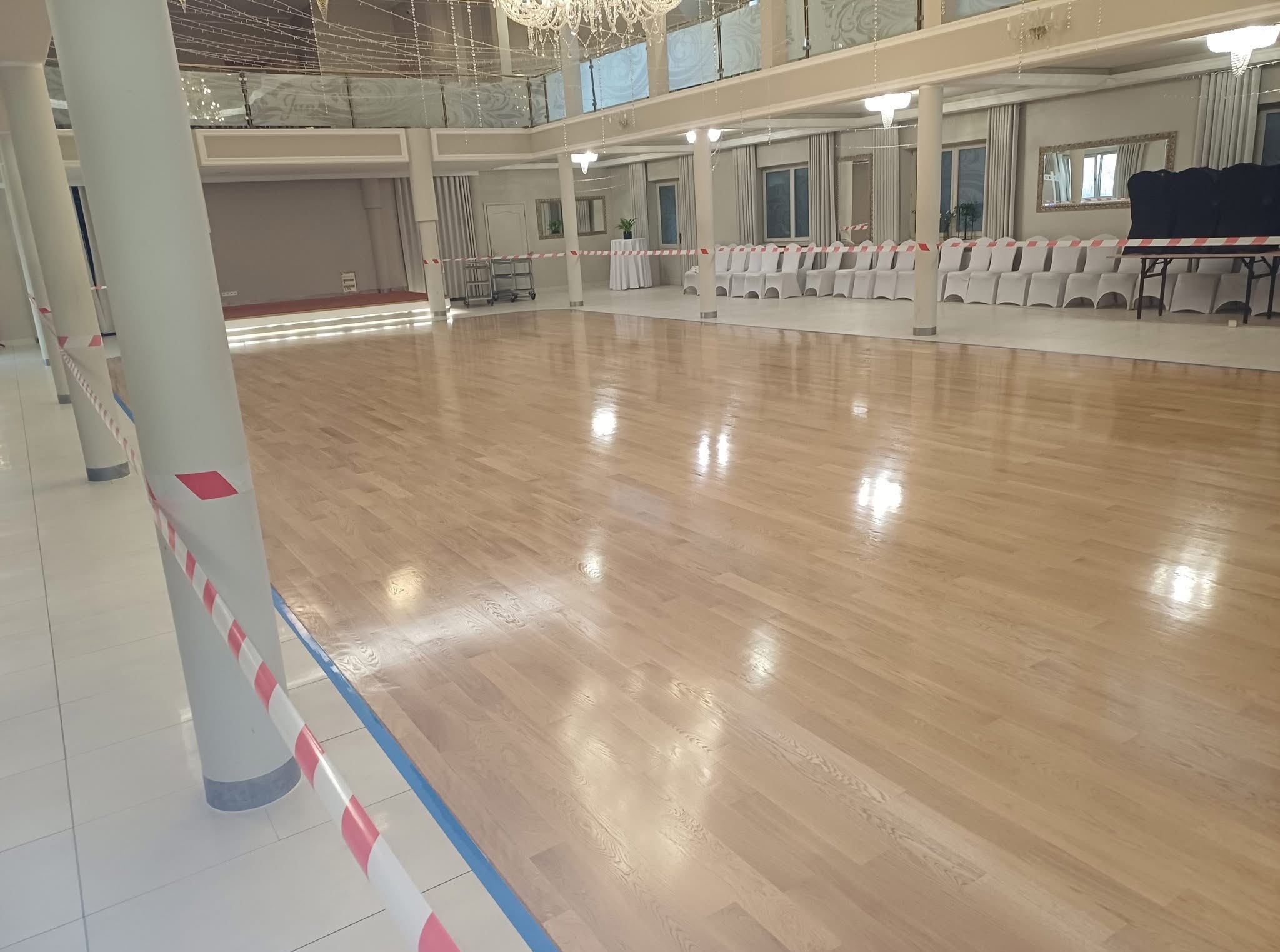 Błyszcząca, jasna podłoga parkietowa w przestronnej sali balowej z kolumnami i dekoracyjnym oświetleniem. Sala przygotowana na wydarzenie, zabezpieczona taśmami.