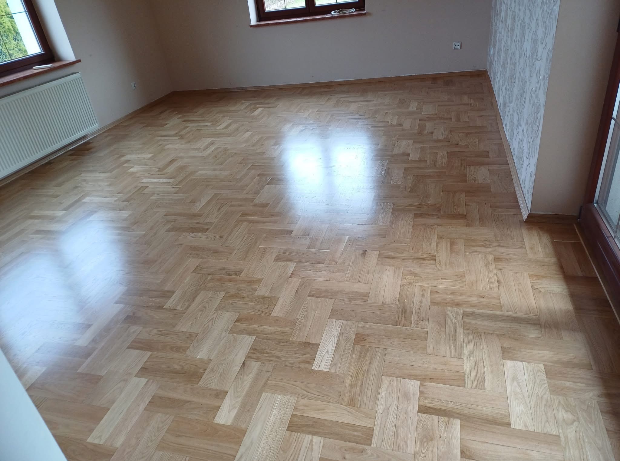 Jasny, parkiet ułożony w jodełkę, po cyklinowaniu, połyskujący w świetle okna, w pustym pokoju z grzejnikiem i oknem.