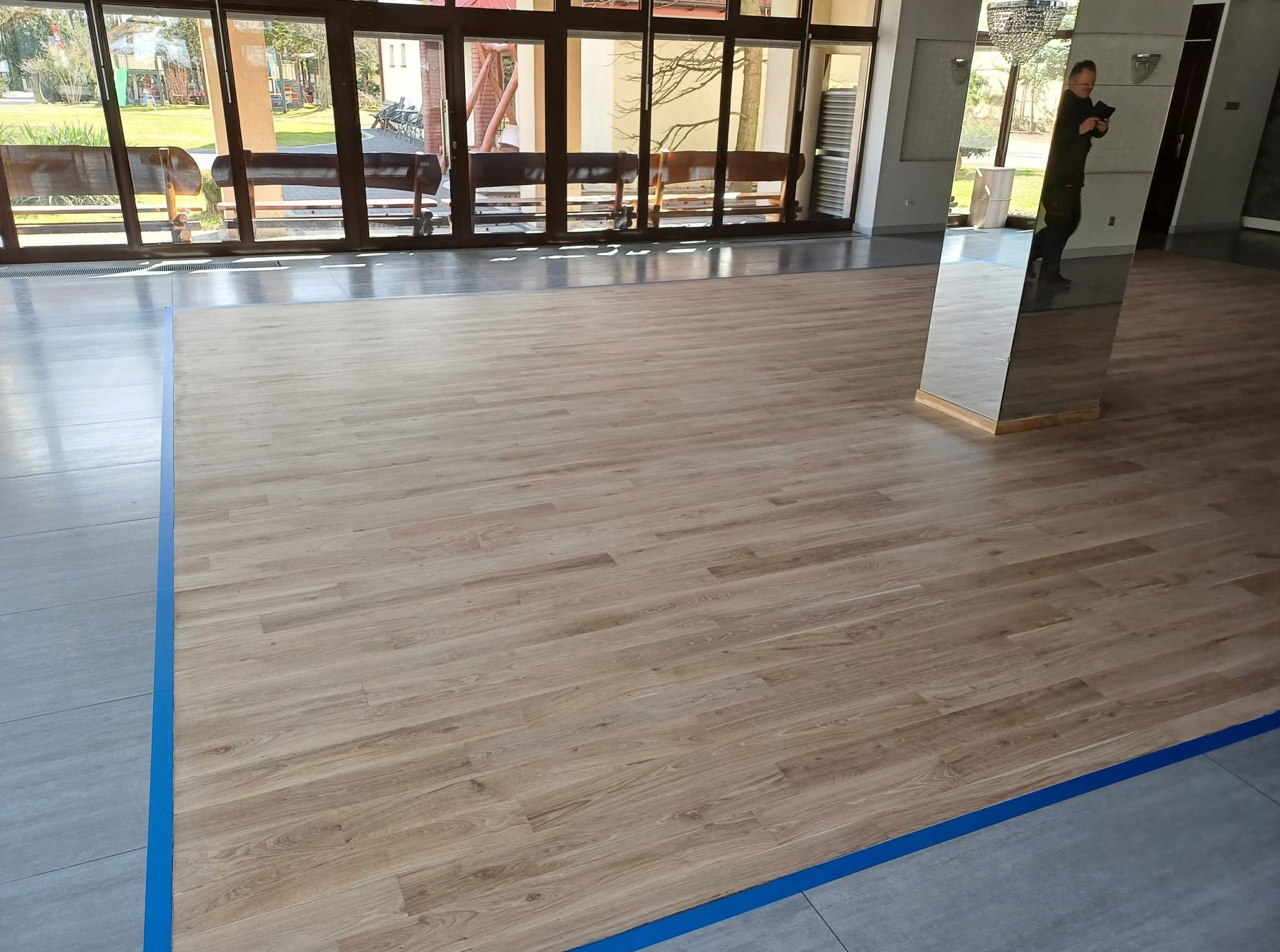 Jasna podłoga z paneli drewnopodobnych, oddzielona niebieską taśmą od szarych płytek. W tle widoczne okna i odbicie osoby w lustrzanej kolumnie.