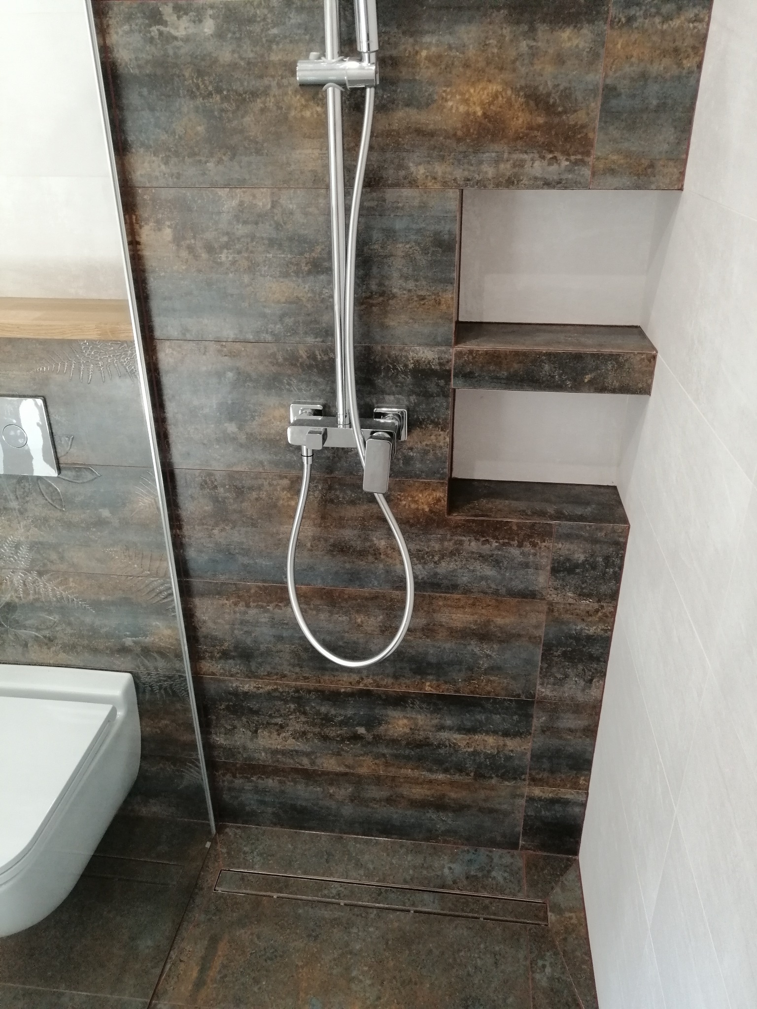 Nowoczesna łazienka z kabiną prysznicową typu walk-in, wykończona płytkami imitującymi drewno i wnęką z półkami. Widoczny odpływ liniowy i minimalistyczna armatura.